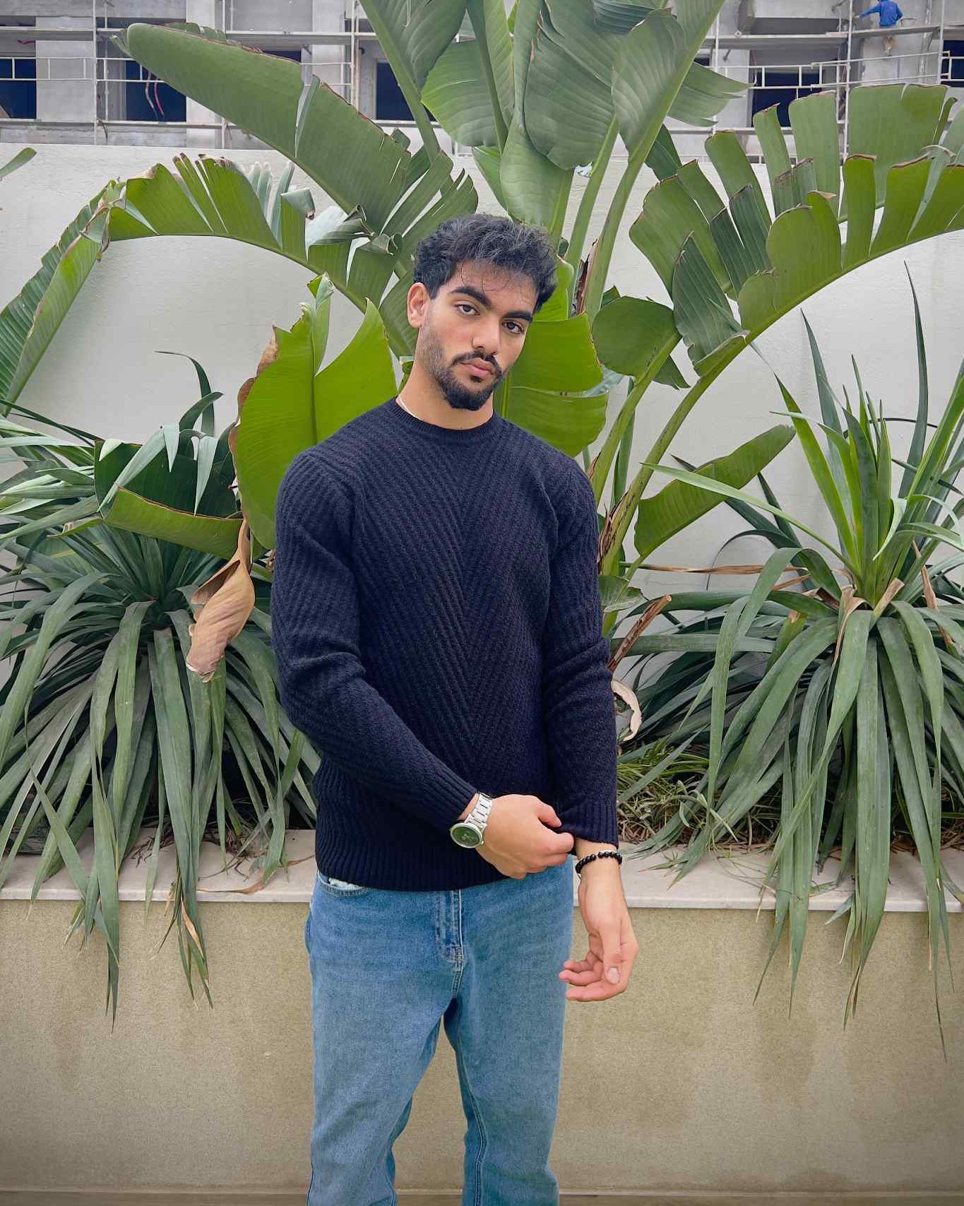 Man wearing a black blue sweater and light blue jeans standing in front of green plants.